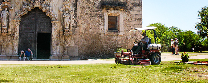 A man operates a commercial-size propane mower in front of a historic mission.