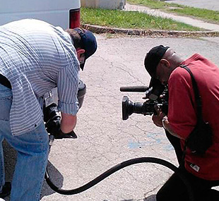 This photo shows a man with a camera filming another man refueling a propane van.