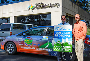A photo of two men posing in front of a vehicle wrapped in graphics that market Clean Cities, holding a sign that says,