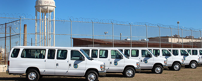 Picture of five white propane-fueled vans in front of prison with barbed wire fence and water tower that says