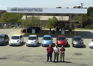 A photo of three men standing in front of seven cars with a fueling station in the background.
