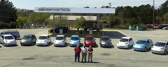 A photo of three men standing in front of seven cars with a fueling station in the background.